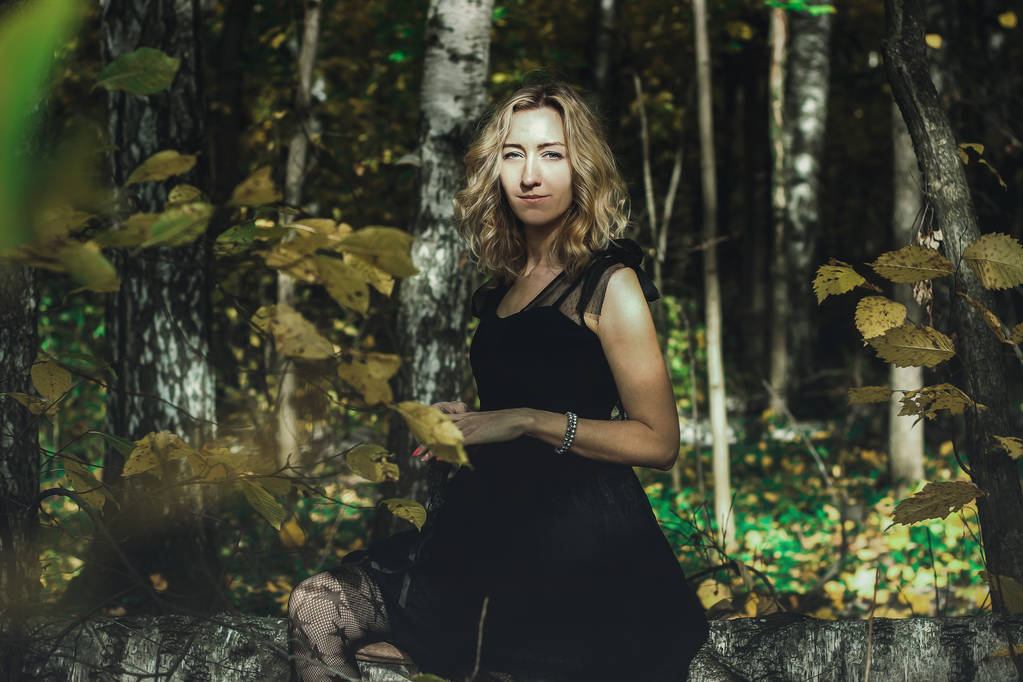 Una bruja joven con un vestido negro con una máscara negra en un bosque místico en varias poses. Transformación artística
. - Foto, imagen