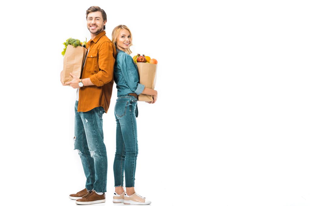 fröhliches Paar mit Einkaufstaschen voller Produkte, die Rücken an Rücken isoliert auf weiß stehen - Foto, Bild