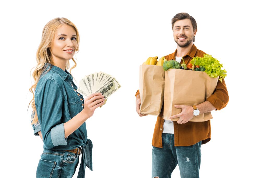 jovem mulher positiva mostrando dinheiro enquanto seu namorado de pé atrás com produtos em sacos de compras isolados no branco
 - Foto, Imagem