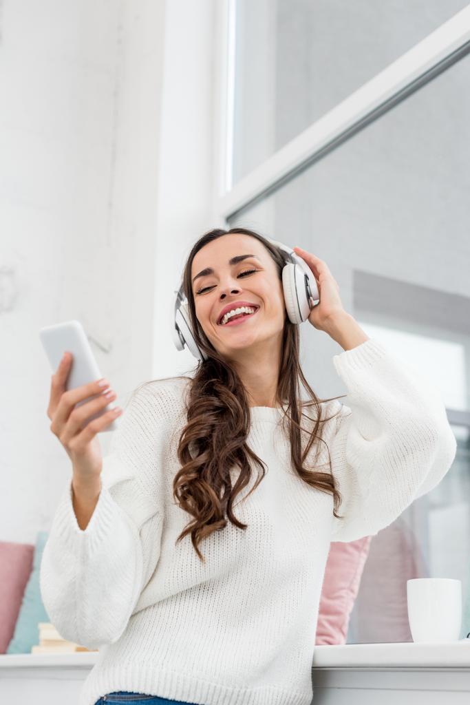 vista inferior de la joven sonriente escuchando música con teléfono inteligente y auriculares inalámbricos en casa
 - Foto, imagen