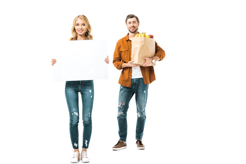 νεαρή γυναίκα δείχνει κενό πανό, ενώ ο φίλος της, στέκεται με προϊόντα σε χάρτινη σακούλα  - Φωτογραφία, εικόνα