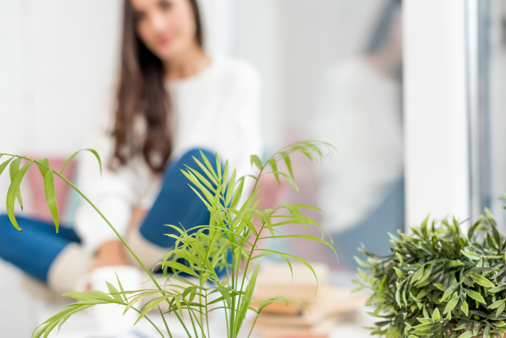 селективное внимание красивой молодой женщины, сидящей дома на подоконнике с комнатными растениями на переднем плане
 - Фото, изображение