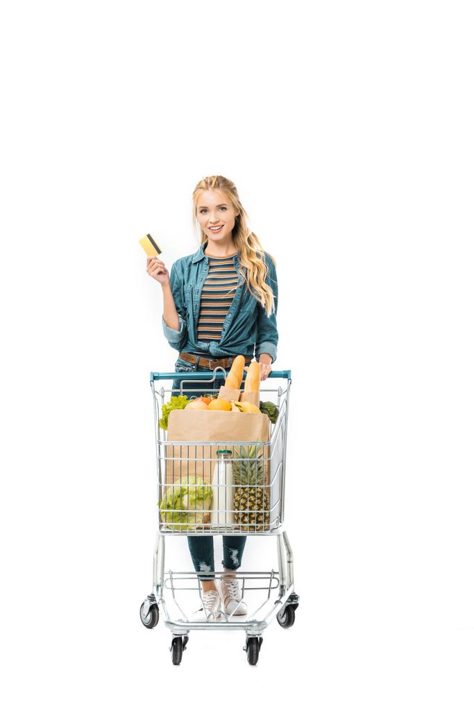 mladá žena ukazuje kreditní kartu a nesoucí nákupní vozík s produkty izolované na bílém - Fotografie, Obrázek