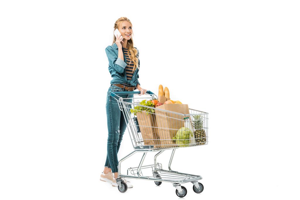 menina atraente falando no smartphone e transportando carrinho de compras com produtos isolados no branco
 - Foto, Imagem