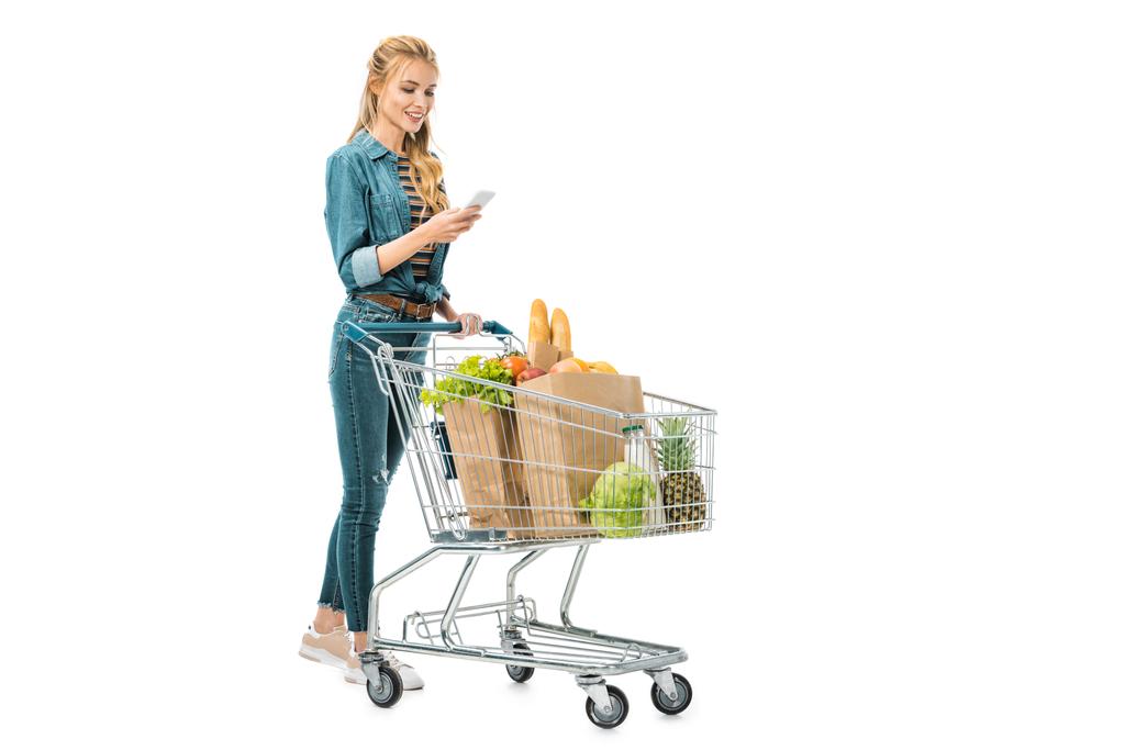 Smartphone kullanarak ve üzerinde beyaz izole ürünleri ile alışveriş arabası taşıyan gülümseyen kadın - Fotoğraf, Görsel