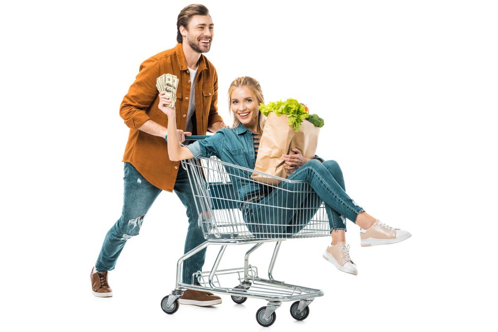schöner Mann mit Einkaufswagen und glücklicher Freundin, die Geld zeigt und Papiertüten mit Produkten auf weißem Papier hält - Foto, Bild