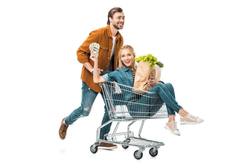 man carrying shopping trolley with happy girlfriend showing money and holding paper bags with products isolated on white - Photo, Image