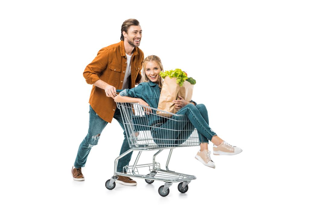 positiver Mann trägt Trolley mit glücklicher Freundin, die Einkaufstüten mit Produkten auf weißem Grund hält - Foto, Bild