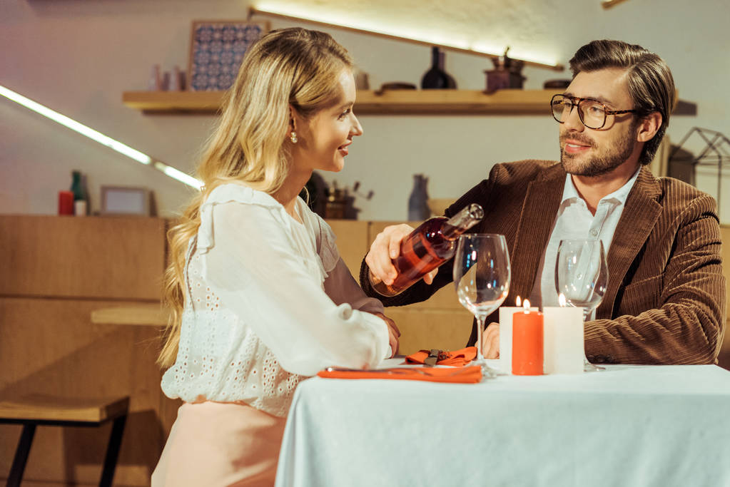 knappe man in jasje gieten wijn in glas aan mooie meisje aan tafel in restaurant  - Foto, afbeelding