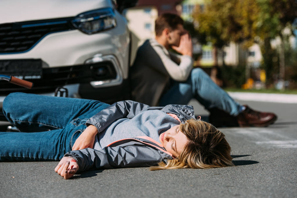 selective focus of injured woman lying on road after car accident with shocked car driver behind  - Photo, Image