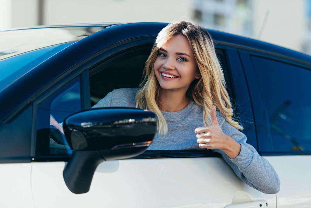 portrait of blond smiling woman showing thumb up while driving car - Photo, Image