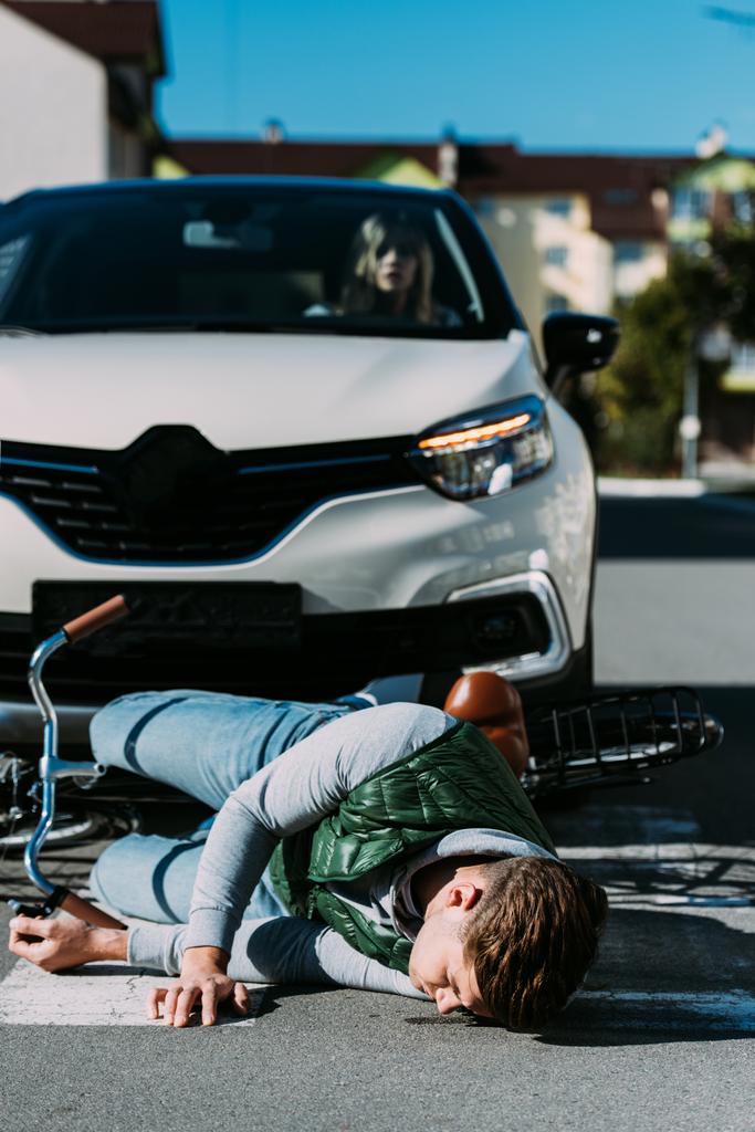жінка сидить у машині і дивиться на пораненого велосипедиста, який лежить на дорозі після зіткнення з дорогою
 - Фото, зображення