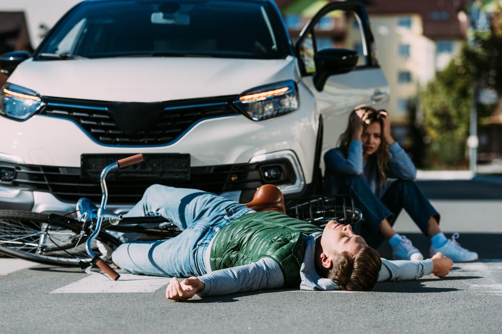 道路と交通衝突後の車の近くに座っている女性の上に横たわる負傷者のサイクリスト - 写真・画像