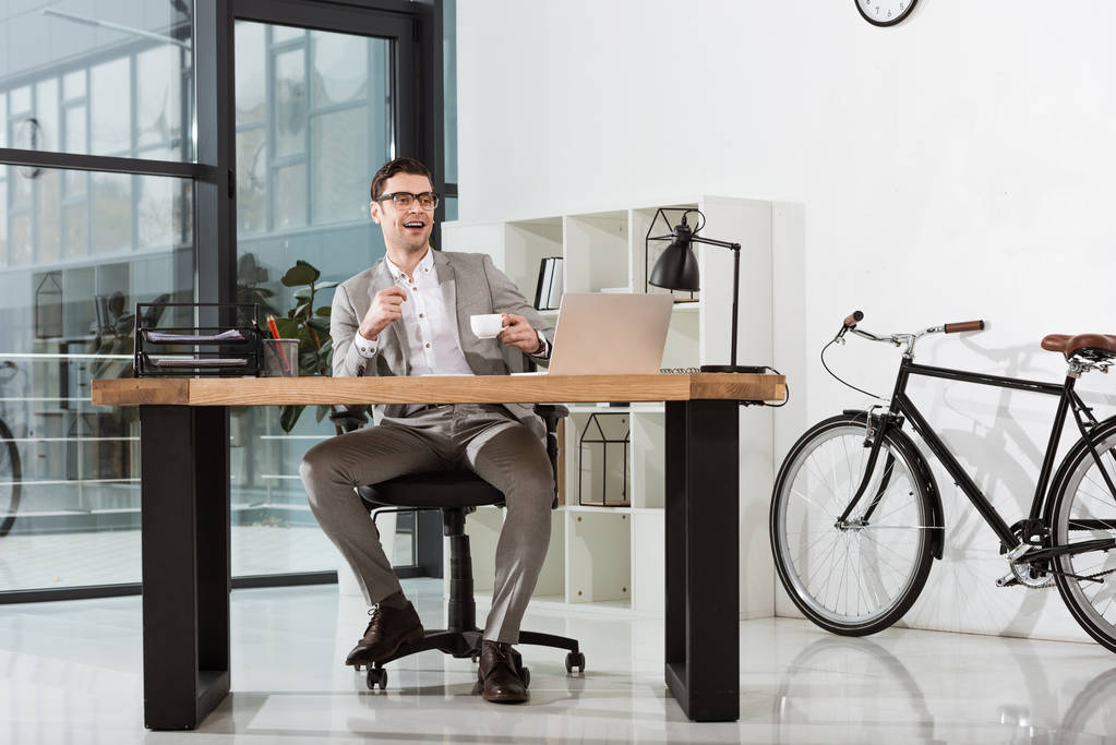 przystojny biznesmen szczęśliwy filiżankę kawy siedząc w pracy w nowoczesnym biurze - Zdjęcie, obraz