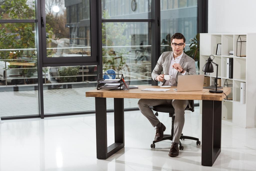 красивый бизнесмен с чашкой кофе сидит на рабочем месте в современном офисе
 - Фото, изображение