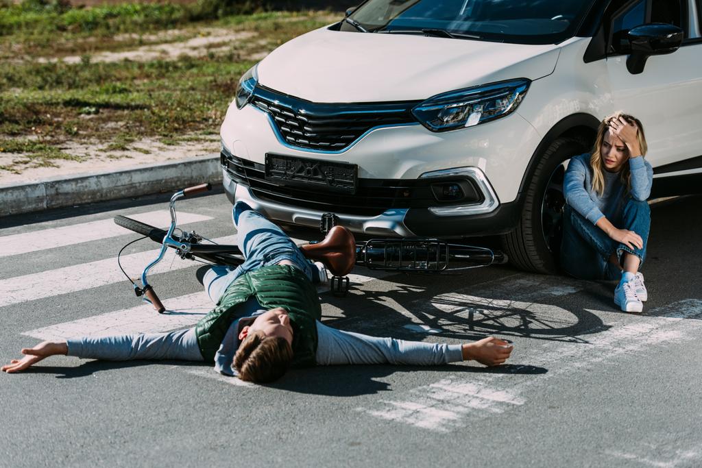 vue grand angle du cycliste couché sur la route et de la femme effrayée pleurant près de la voiture après une collision de la circulation
  - Photo, image