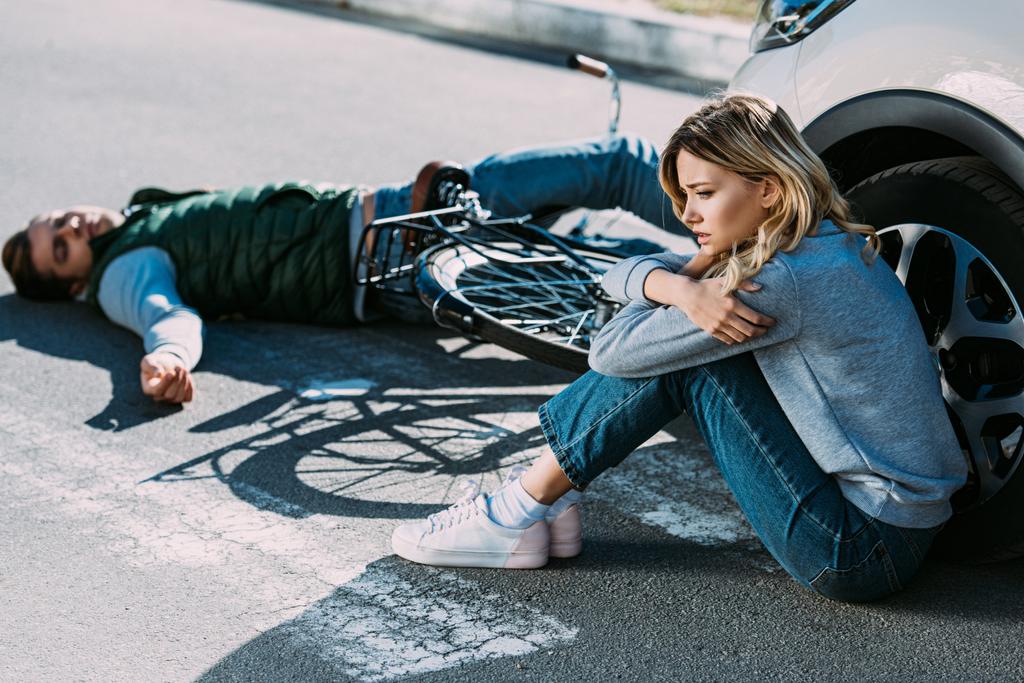 hoge hoekmening van jonge vrouw zitten in de buurt van de auto en slachtoffer van het auto-ongeluk op weg liggen - Foto, afbeelding