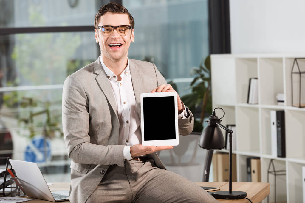 beau homme d'affaires heureux assis sur le bureau sur le lieu de travail et tenant tablette avec écran blanc
 - Photo, image