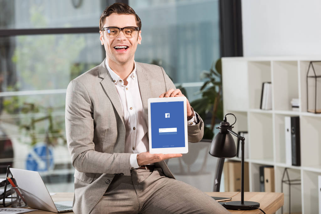 handsome happy businessman sitting on desk at workplace and holding tablet with facebook website on screen - Photo, Image