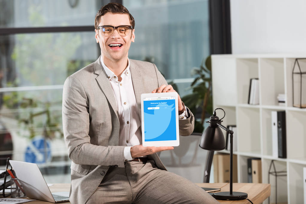 schöner glücklicher Geschäftsmann, der am Schreibtisch am Arbeitsplatz sitzt und Tablet mit Twitter-Webseite auf dem Bildschirm hält - Foto, Bild