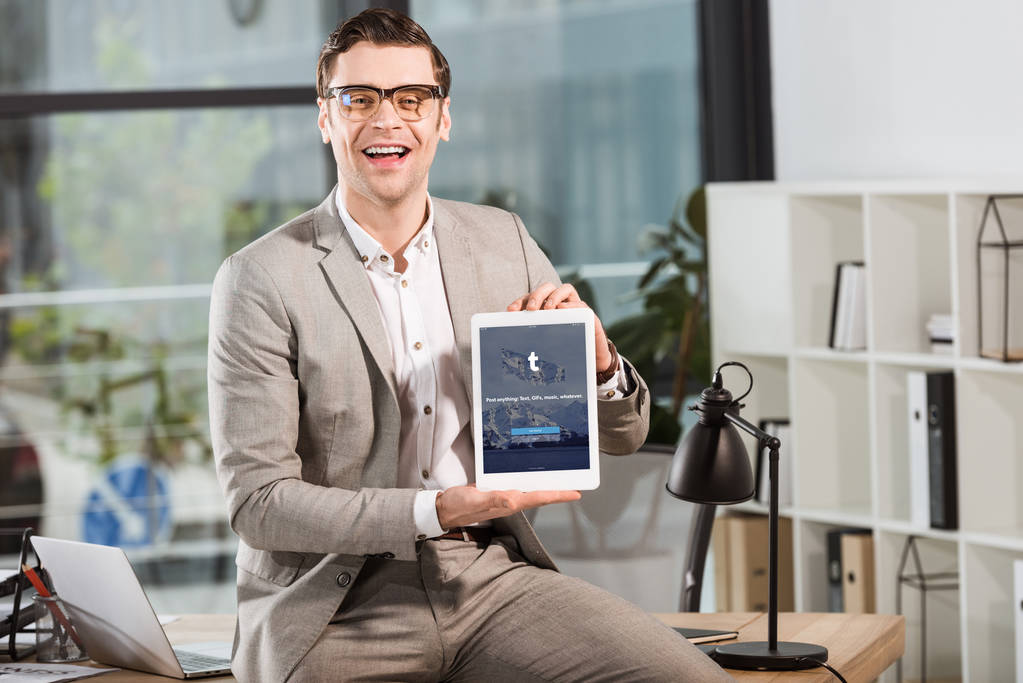handsome happy businessman sitting on desk at workplace and holding tablet with tumblr website on screen - Photo, Image