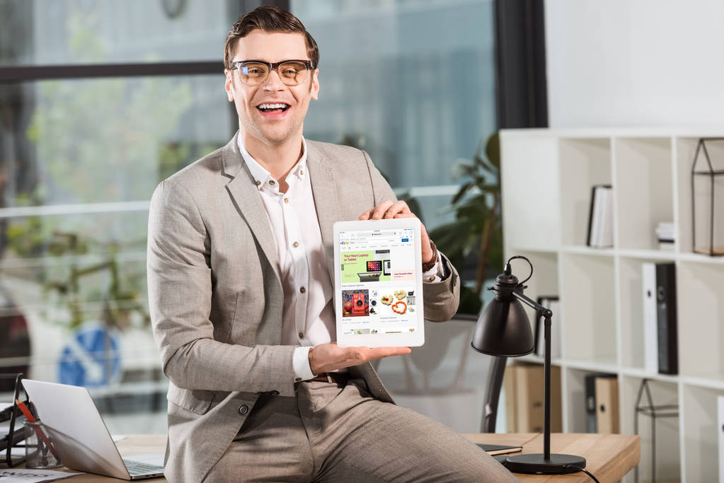 красивый счастливый бизнесмен, сидящий за рабочим столом и держащий в руках табличку с сайтом Ebay на экране
 - Фото, изображение