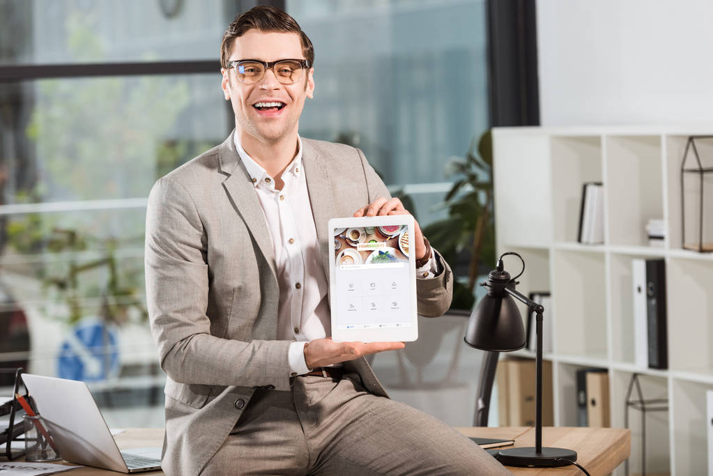 handsome happy businessman sitting on desk at workplace and holding tablet with foursquare website on screen - Photo, Image