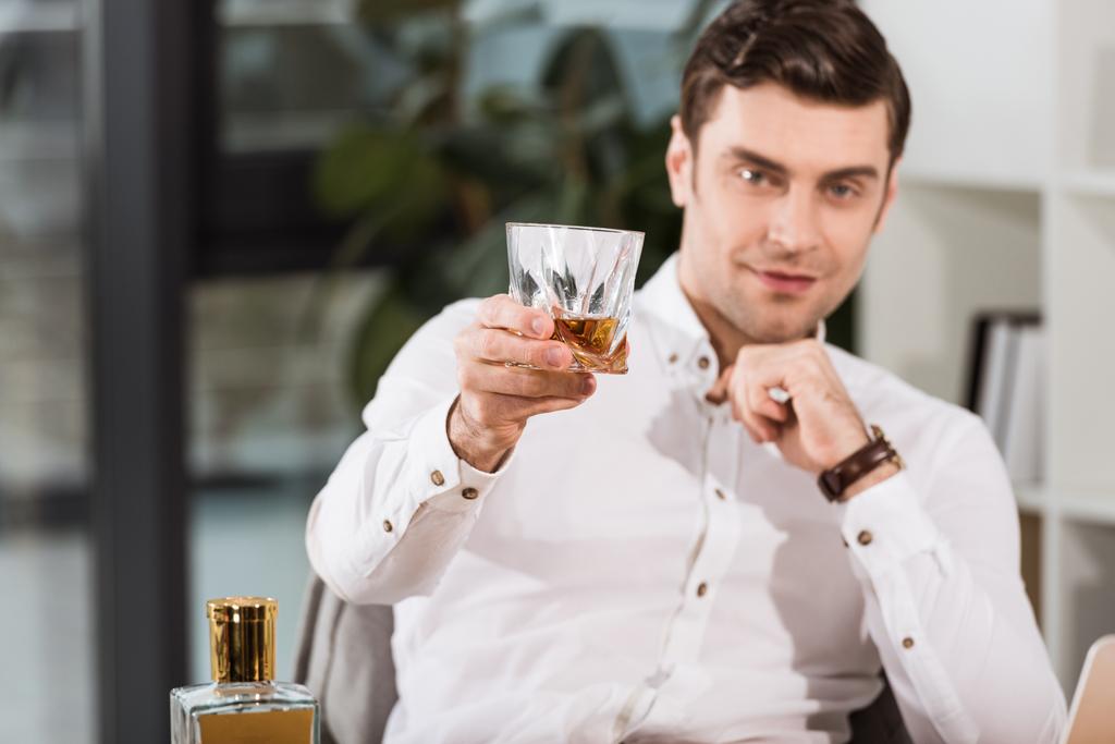 handsome businessman holding glass of whiskey and looking at camera at office - Photo, Image