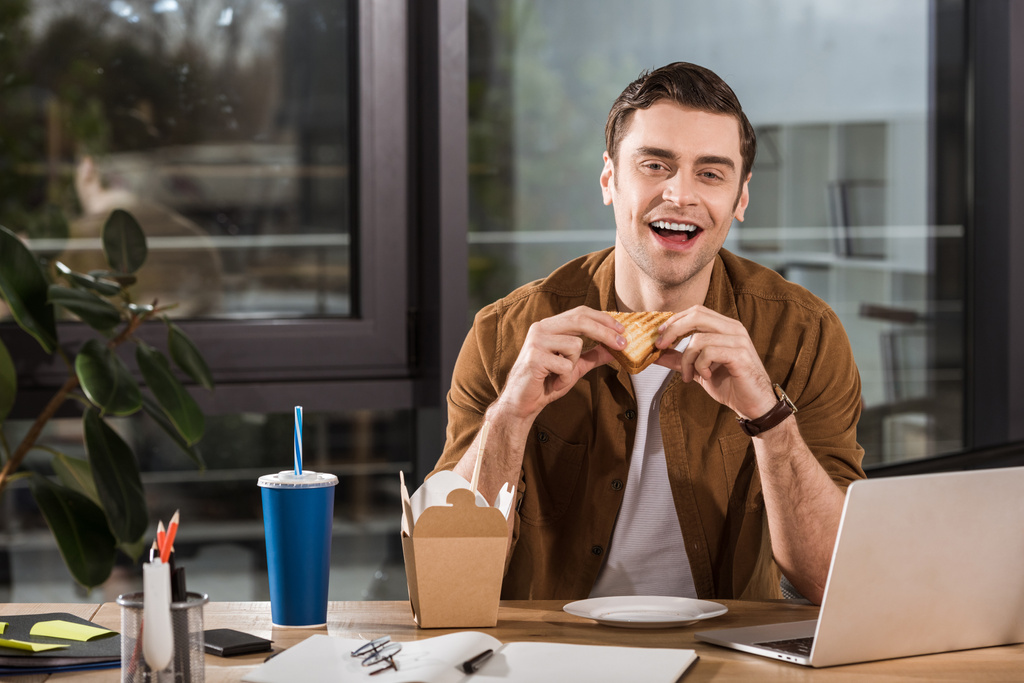 guapo feliz hombre de negocios comiendo sándwich y llevar fideos en la oficina
 - Foto, imagen
