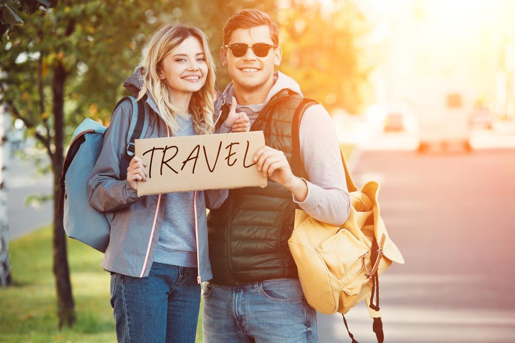 feliz joven pareja de viajeros con tarjeta de viaje con inscripción y sonriendo a la cámara en la carretera
 - Foto, imagen