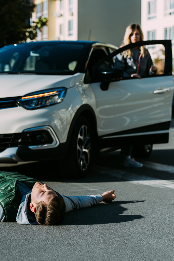 joven herido acostado en la carretera y la mujer de pie cerca de coche detrás después de la colisión de tráfico
 - Foto, Imagen