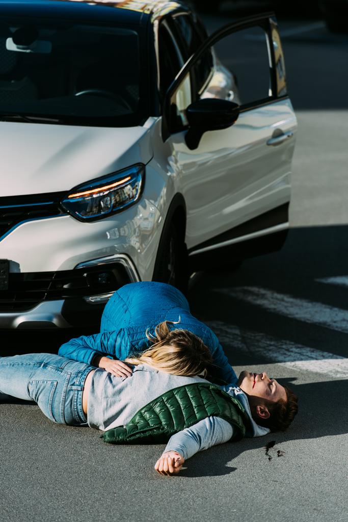 vue en angle élevé de la femme vérifiant le rythme cardiaque de l'homme blessé allongé sur la route après un accident de voiture
  - Photo, image