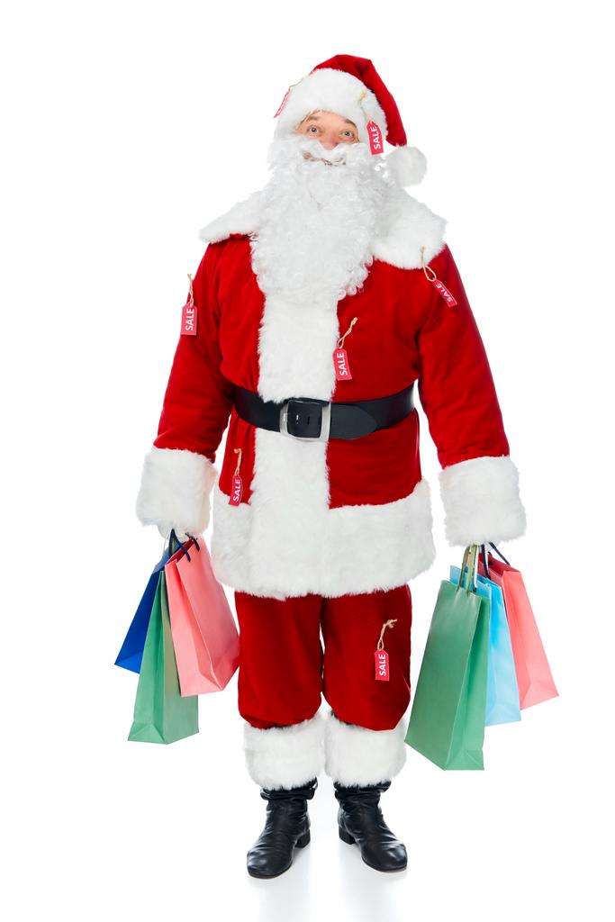 happy santa claus in red costume with sale tags holding shopping bags isolated on white - Photo, Image