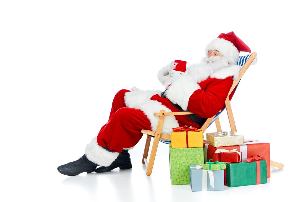 Père Noël claus tenant boisson chaude et assis sur une chaise de plage avec des cadeaux de Noël sur blanc
 - Photo, image