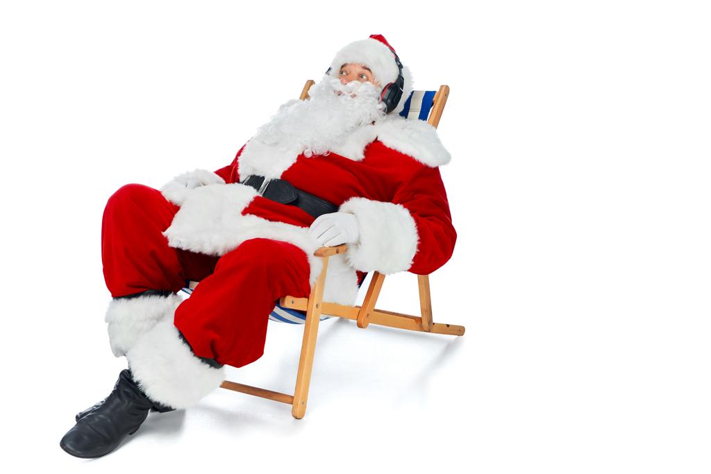 Санта Клаус слушает музыку в наушниках, отдыхая на пляжном стуле на белом
 - Фото, изображение