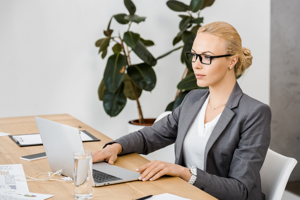 young woman in glasses sitting at table and using laptop in insurance office - Photo, Image