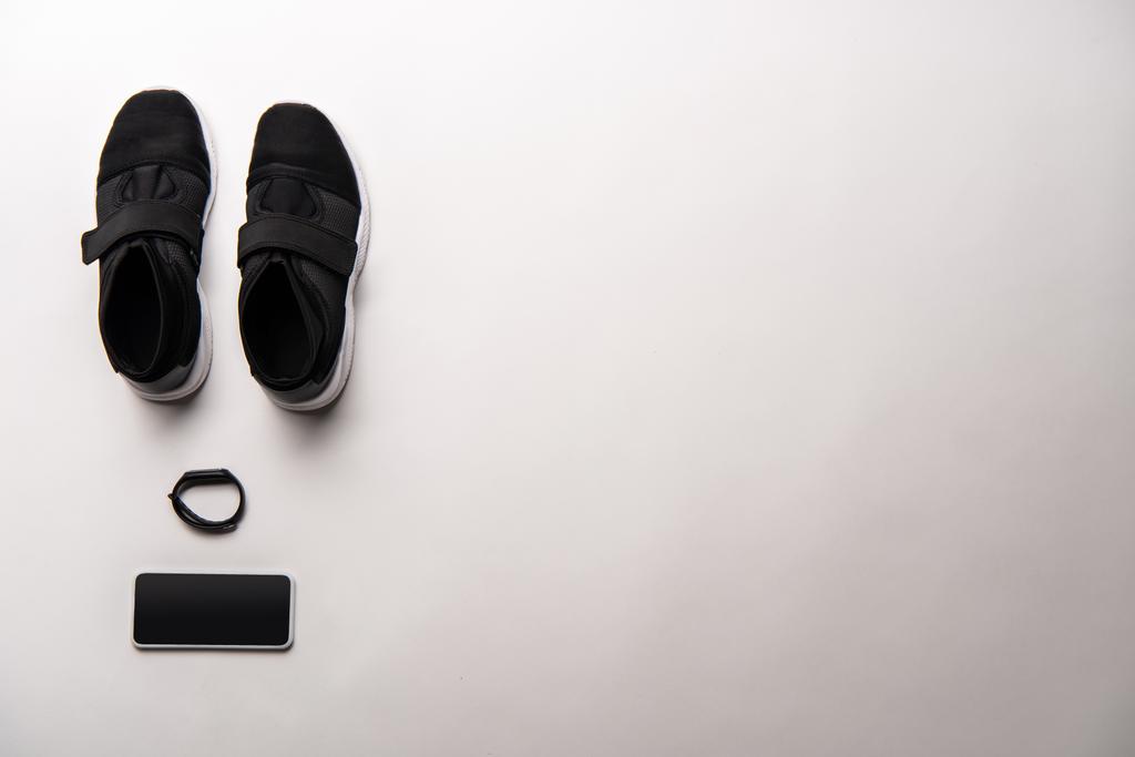 θέα από ψηλά τοποθετημένα μαύρα αθλητικά παπούτσια, fitness tracker και smartphone με κενή οθόνη που απομονώνονται σε λευκό - Φωτογραφία, εικόνα