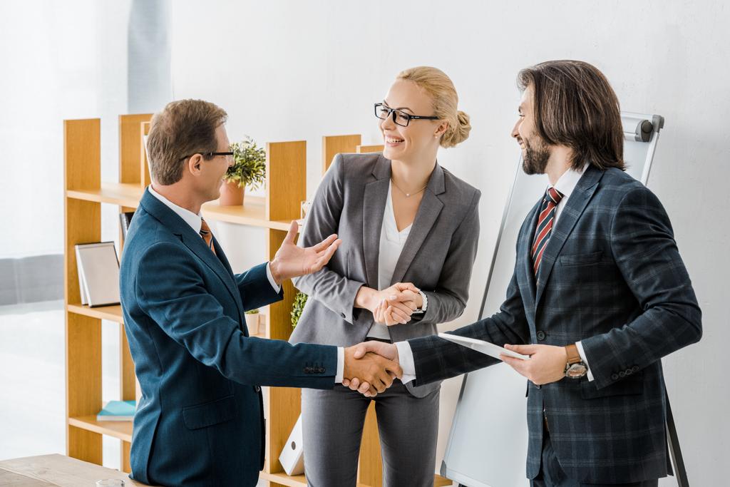 two businessmen shaking hands and talking to smiling businesswoman in office - Photo, Image