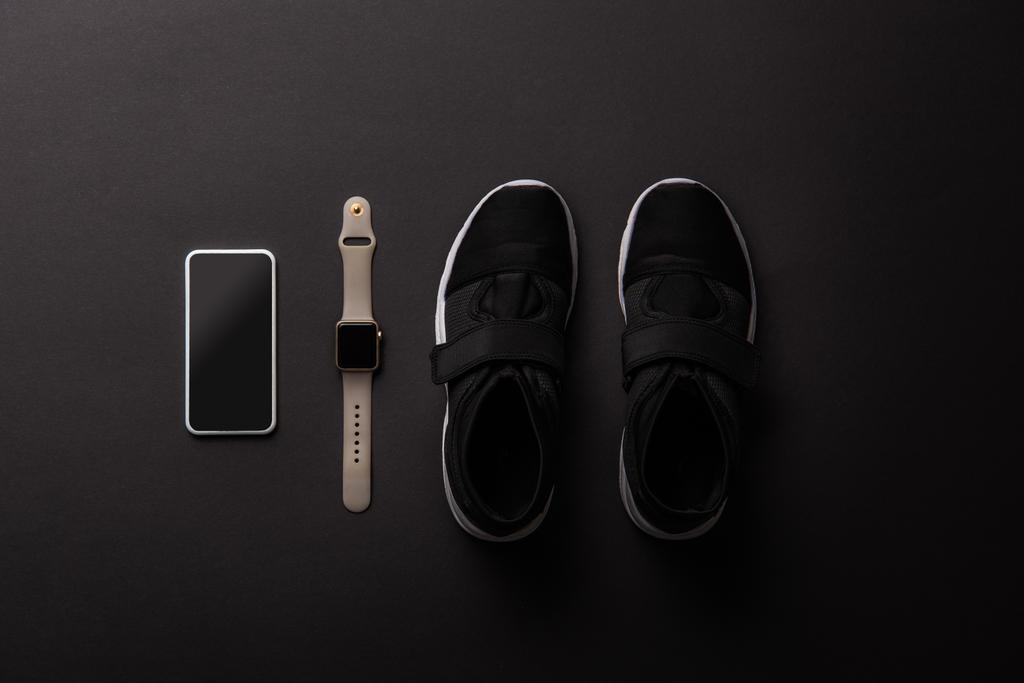 verhoogde oogpunt geschikt smartphone met leeg scherm, smartwatch en sneakers geïsoleerd op zwart - Foto, afbeelding