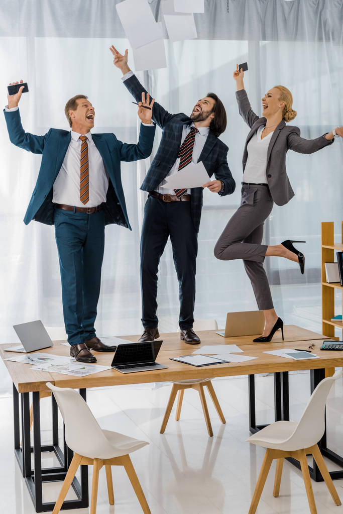 joyful insurance workers dancing on table and throwing papers at meeting in office - Photo, Image