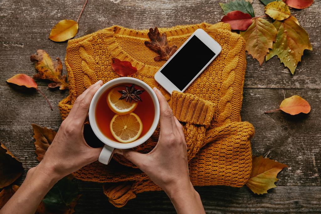 частковий вигляд жінки, що тримає чашку чаю на дерев'яній стільниці з в'язаним светром, смартфоном і опалим листям
 - Фото, зображення