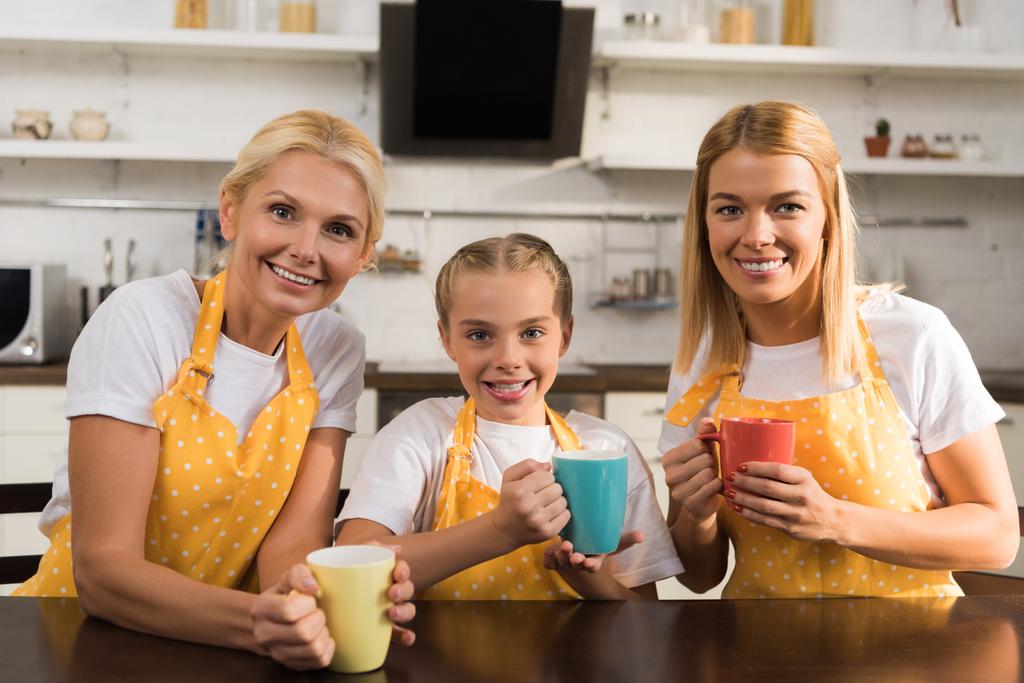 красива щаслива сім'я трьох поколінь в фартухах, що тримає барвисті чашки і посміхається на камеру
 - Фото, зображення