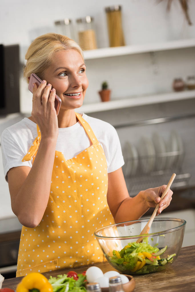 усміхнена зріла жінка в фартусі розмовляє по смартфону і дивиться в сторону під час приготування овочевого салату на кухні
 - Фото, зображення