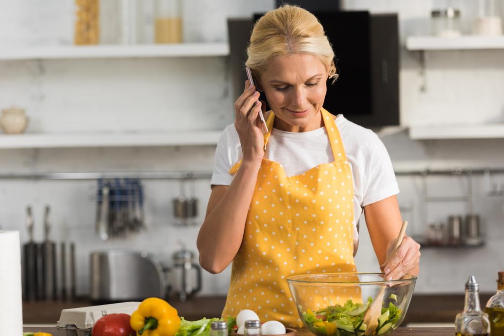 χαμογελώντας ώριμη γυναίκα στην ποδιά μιλάμε από smartphone, ενώ το μαγείρεμα σαλάτα λαχανικών  - Φωτογραφία, εικόνα