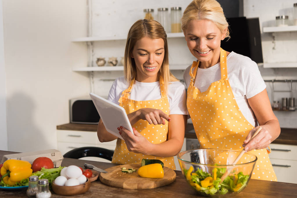 щаслива зріла жінка з дорослою дочкою використовує цифровий планшет під час приготування їжі разом
  - Фото, зображення