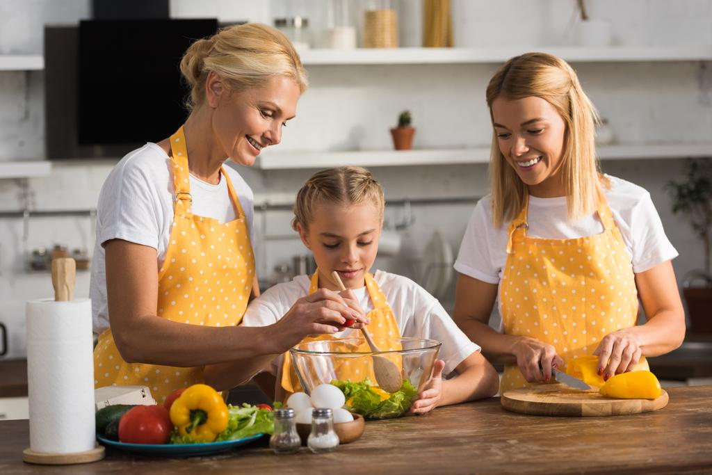 glückliches Kind mit Mutter und Großmutter beim gemeinsamen Kochen von Gemüsesalat in der Küche  - Foto, Bild