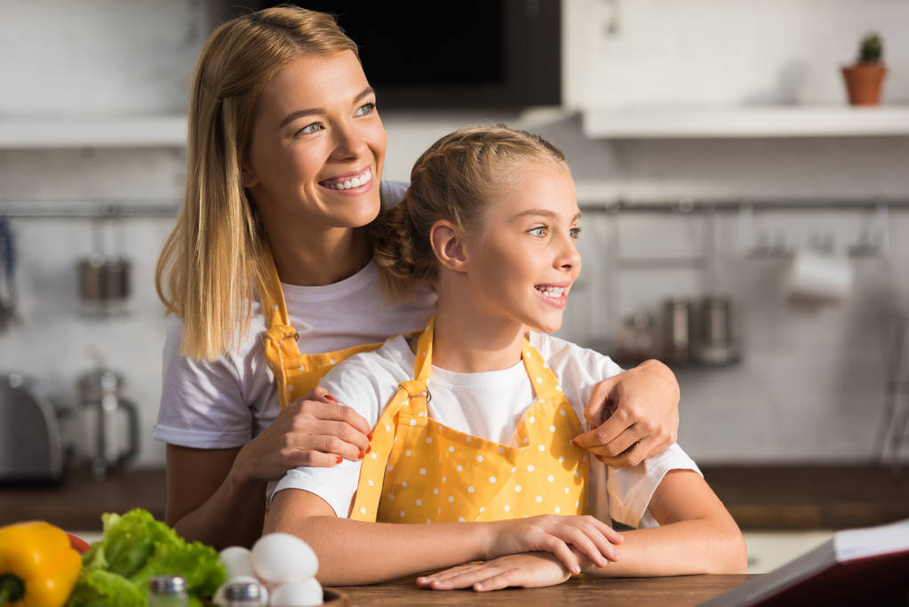 feliz madre e hija en delantales sonriendo y mirando hacia otro lado en la cocina
 - Foto, imagen