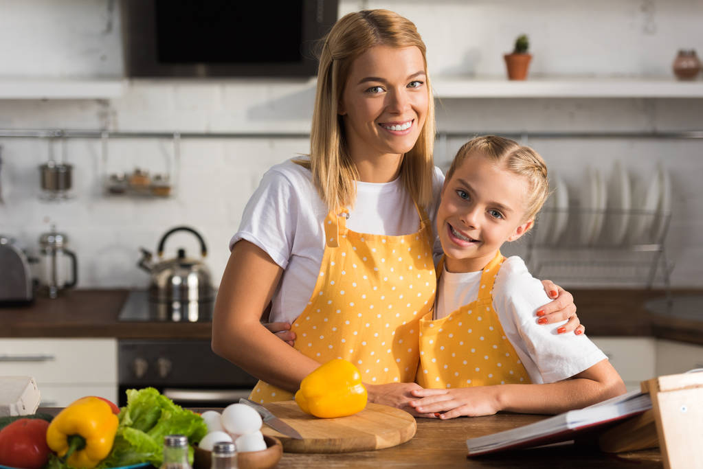 bela mãe e filha feliz em aventais sorrindo para a câmera enquanto cozinhando juntos
  - Foto, Imagem