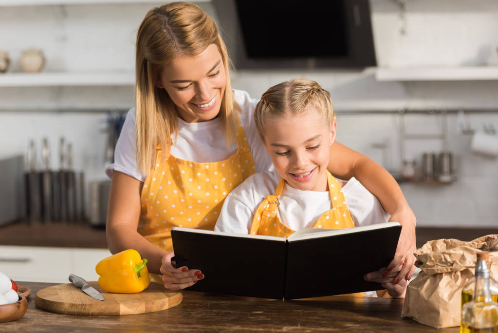 happy mother and daughter reading cookbook while cooking together - Photo, Image