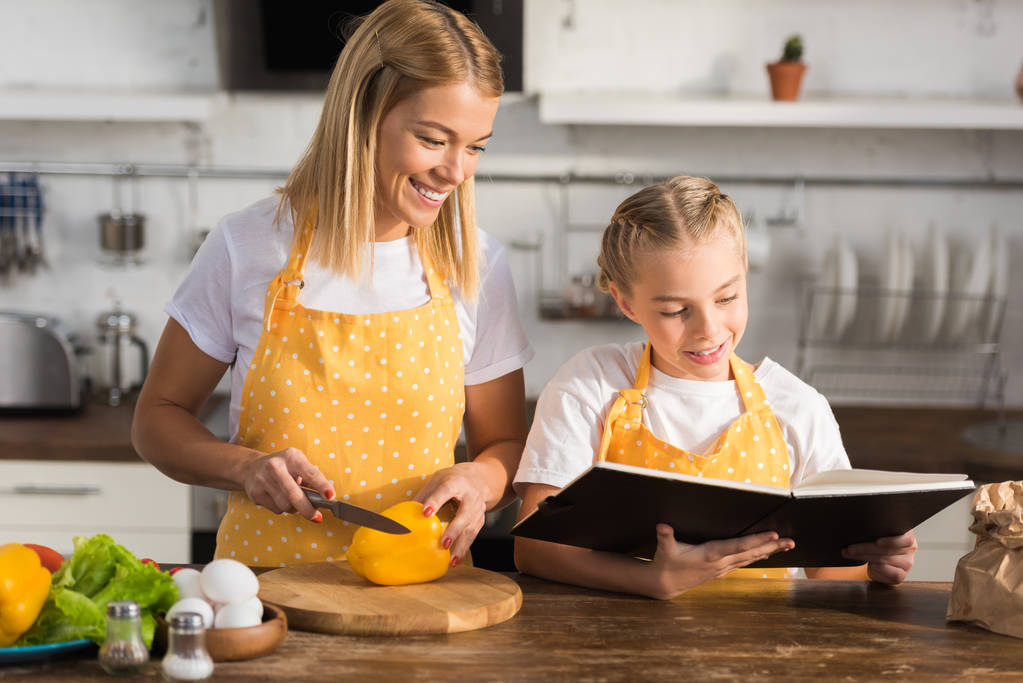 souriant jeune mère couper le poivre et regarder fille mignonne lecture livre de cuisine
 - Photo, image